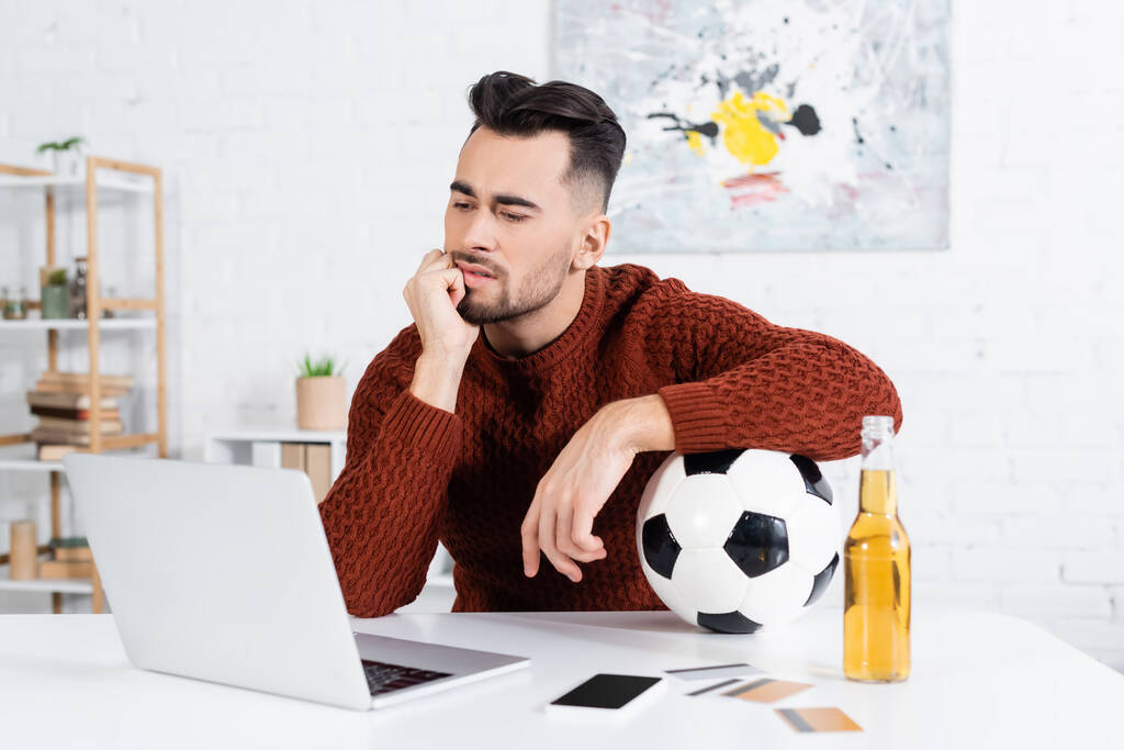 thoughtful bookmaker with soccer ball looking at laptop near smartphone and credit cards - Foto, Imagen