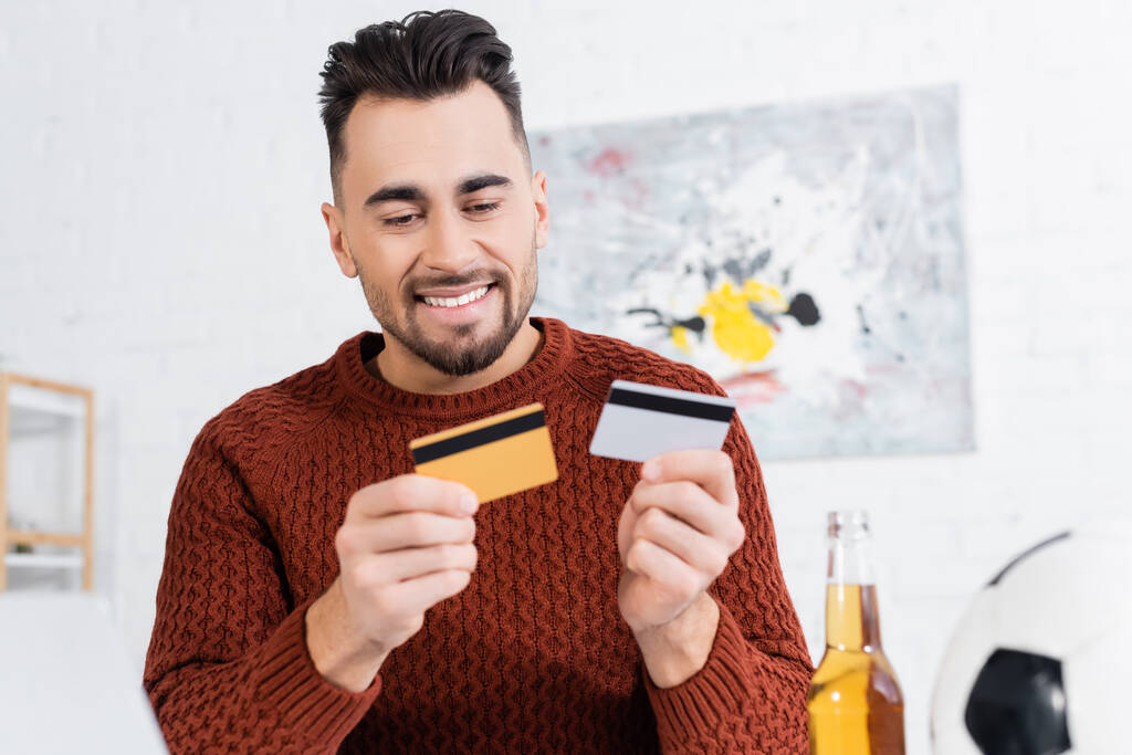 casa de apuestas sonriente sosteniendo tarjetas de crédito cerca borrosa pelota de fútbol y cerveza - Foto, Imagen