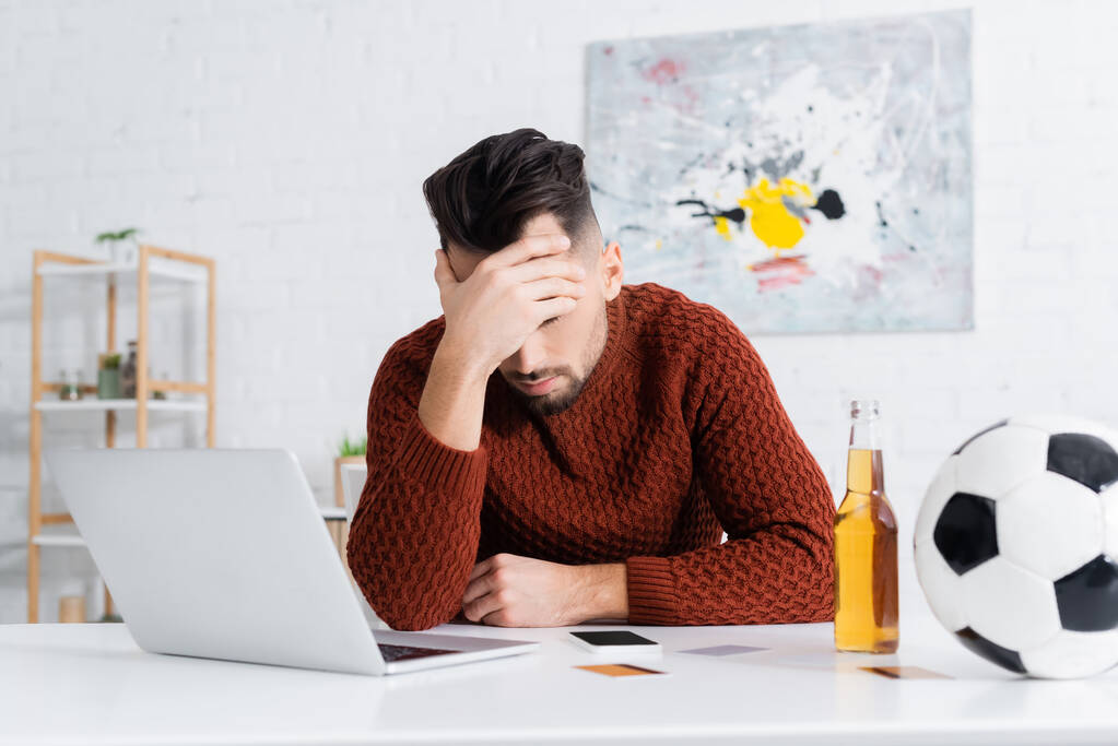 depressieve bookmaker zit met gebogen hoofd in de buurt van laptop, voetbal en fles bier - Foto, afbeelding