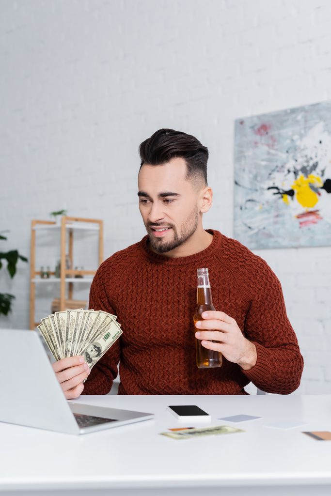 giocatore d'azzardo sorridente con dollari e bottiglia di birra vicino a laptop e smartphone con schermo bianco - Foto, immagini