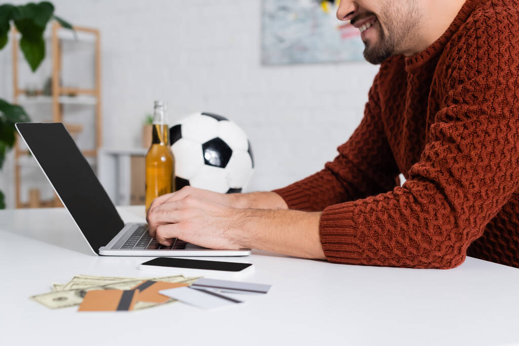 vista ritagliata di bookmaker sorridente digitando sul computer portatile vicino a carte di credito e pallone da calcio - Foto, immagini