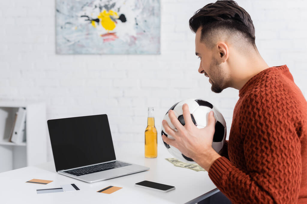 vista laterale del giocatore d'azzardo con pallone da calcio vicino al computer portatile con schermo bianco, carte di credito e bottiglia di birra - Foto, immagini