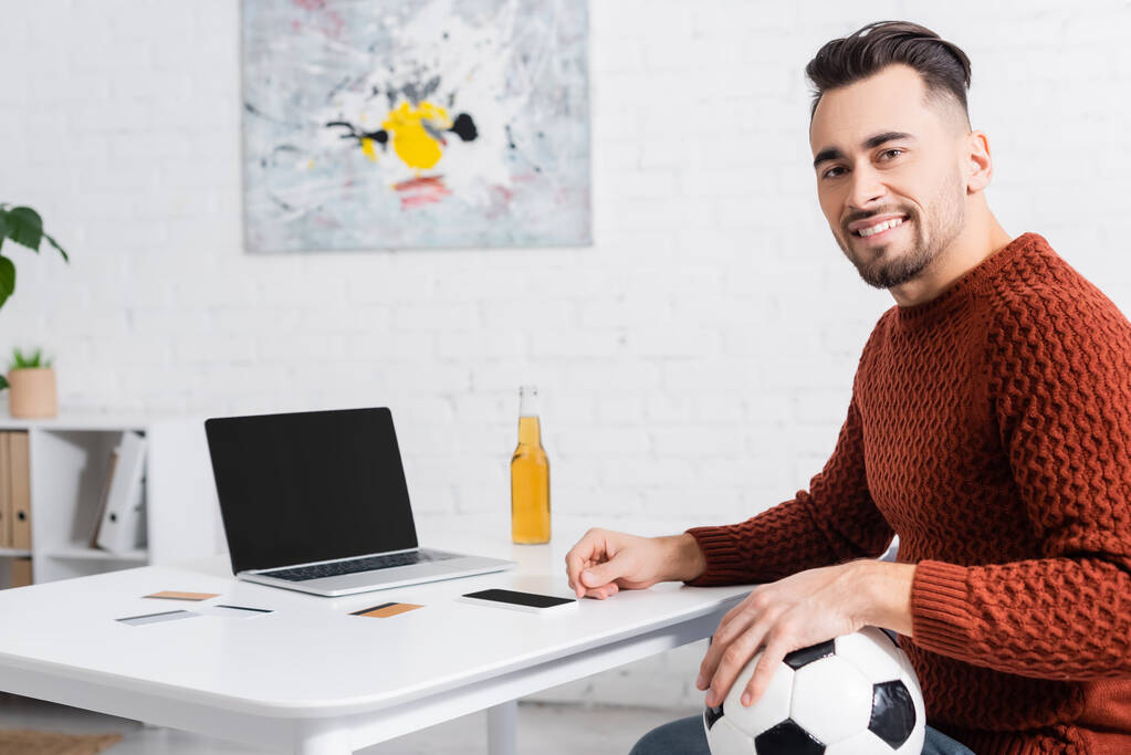 šťastný hráč s fotbalovým míčem při pohledu do kamery v blízkosti notebooku a kreditních karet - Fotografie, Obrázek