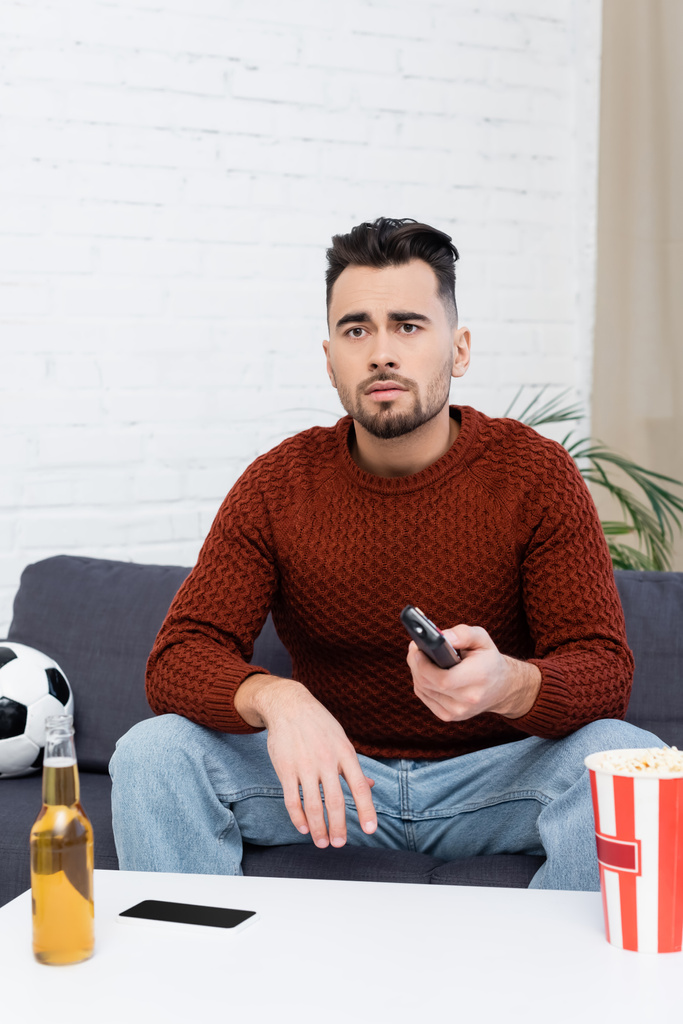 smutný sportovní fanoušek s dálkovým ovladačem sledování hry v televizi v blízkosti fotbalového míče, piva a popcornu - Fotografie, Obrázek