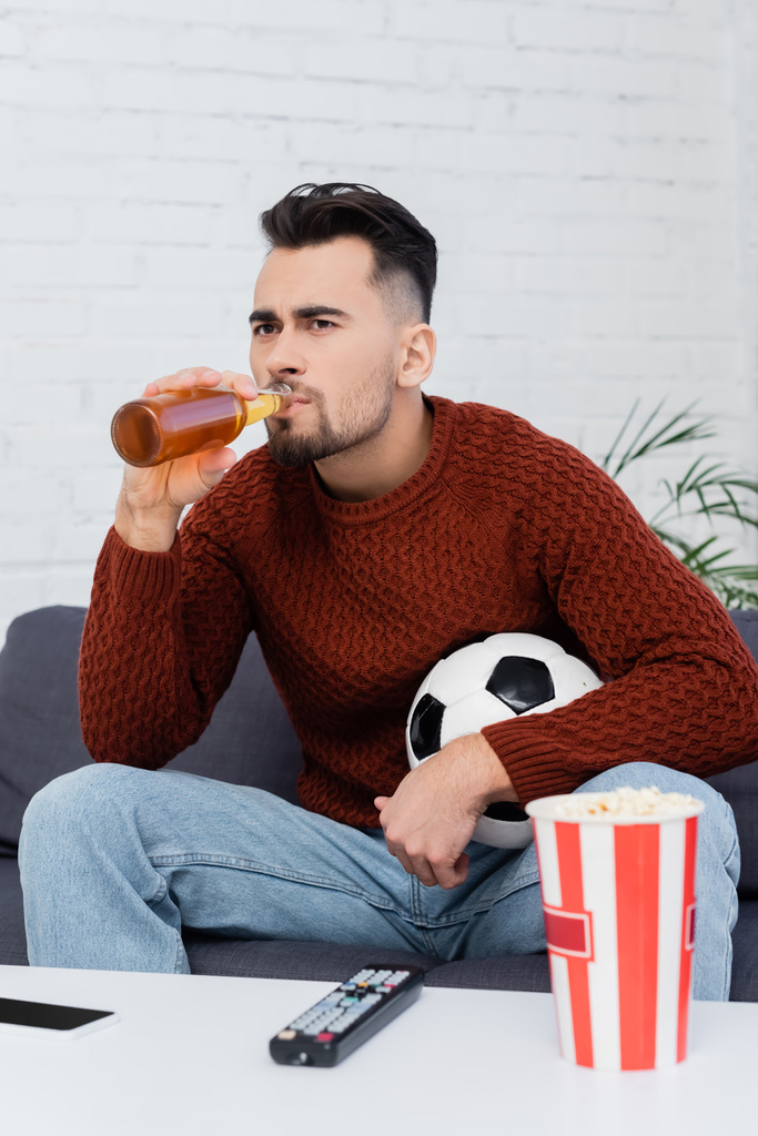 fanático del fútbol enfocado beber cerveza mientras ve el juego en la televisión en casa - Foto, Imagen