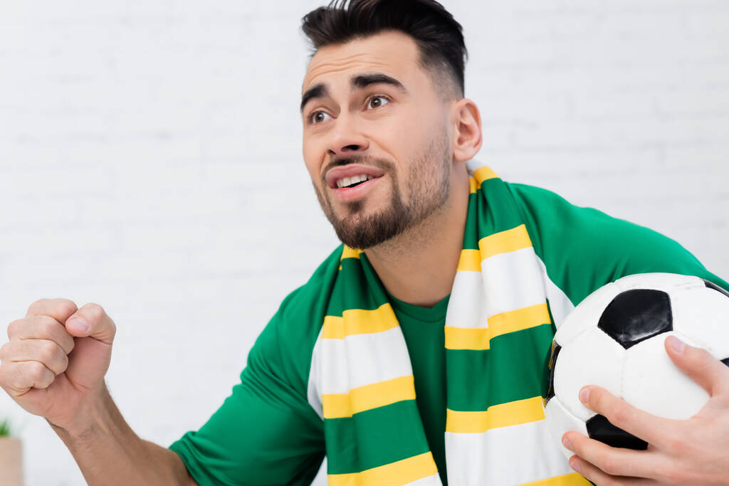 возбужденный спортивный болельщик держит футбольный мяч и сжимает кулак на удачу во время просмотра чемпионата - Фото, изображение