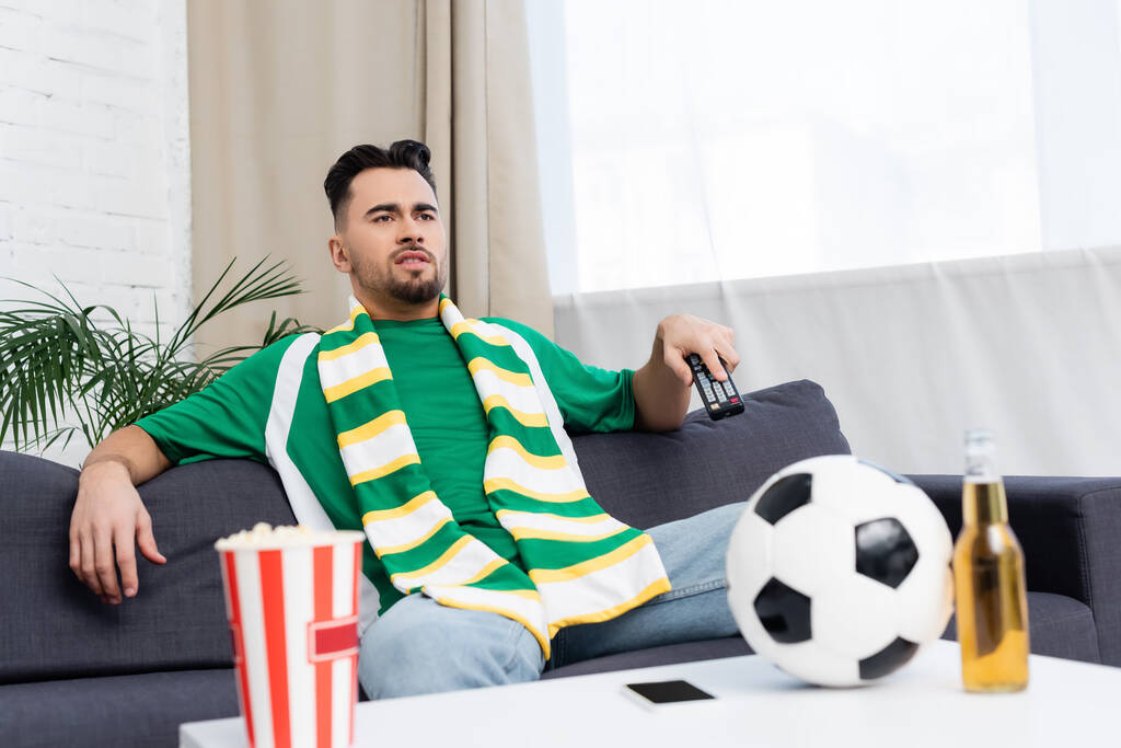 sports fan in striped scarf watching game on tv near blurred soccer ball, beer and popcorn - Fotó, kép