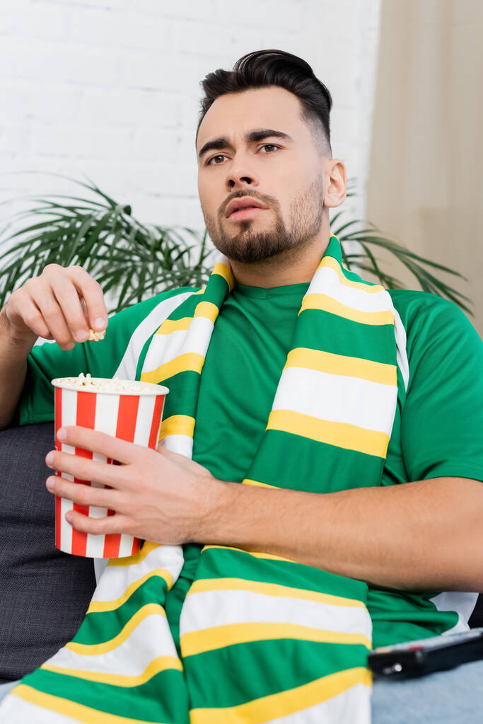 hombre enfocado en bufanda rayada sosteniendo un cubo de papel de palomitas de maíz y viendo el juego deportivo en casa - Foto, imagen