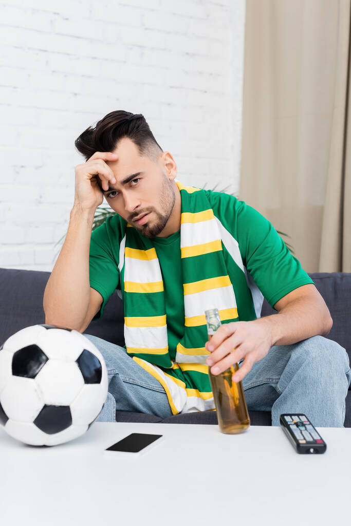 frustrated sports fan with bottle of beer watching match on tv near soccer ball - Фото, изображение