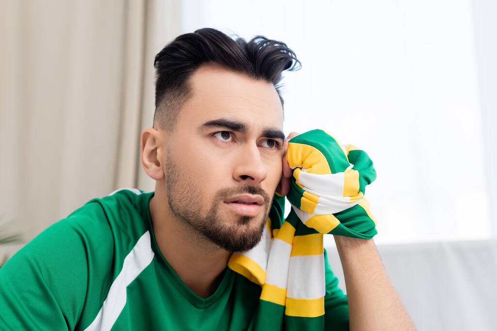 расстроенный мужчина с полосатым шарфом смотрит спортивный матч дома - Фото, изображение