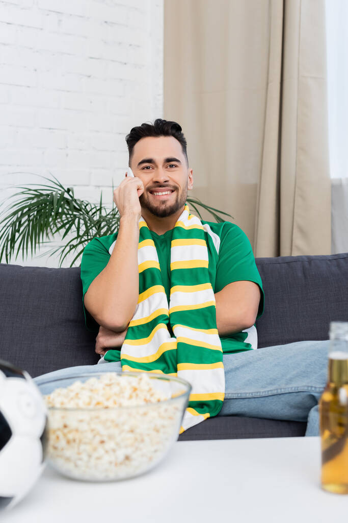 Счастливый футбольный болельщик разговаривает по смартфону рядом с размытой кукурузой и пивом - Фото, изображение