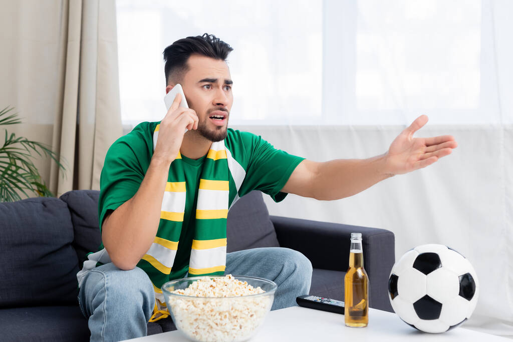 displeased sports fan pointing with hand and talking on smartphone while watching football match on tv - Photo, image