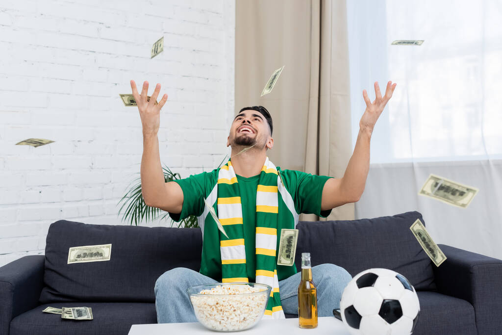excited bookmaker throwing up money near soccer ball, beer and popcorn - Foto, afbeelding