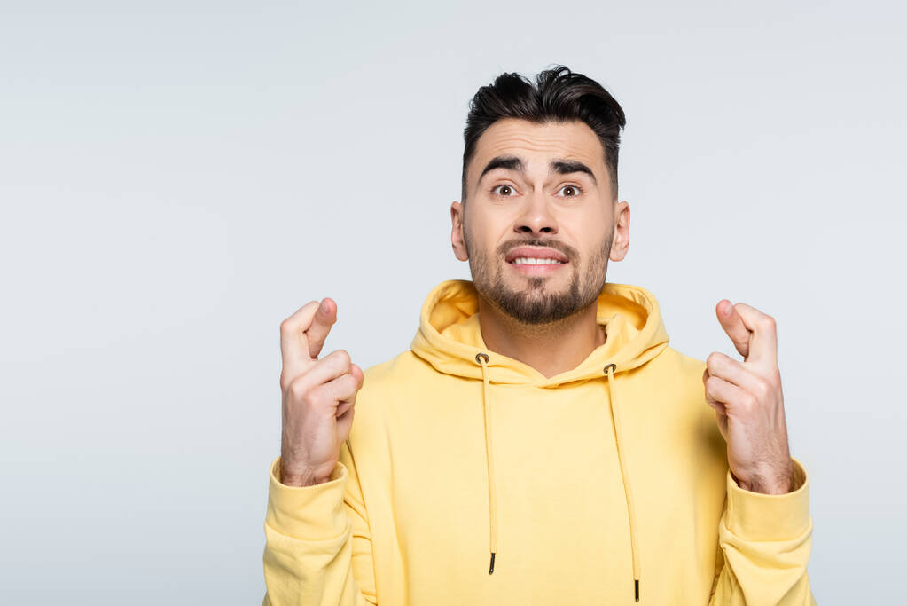 worried gambler in yellow hoodie holding crossed fingers for luck isolated on grey - Foto, Bild