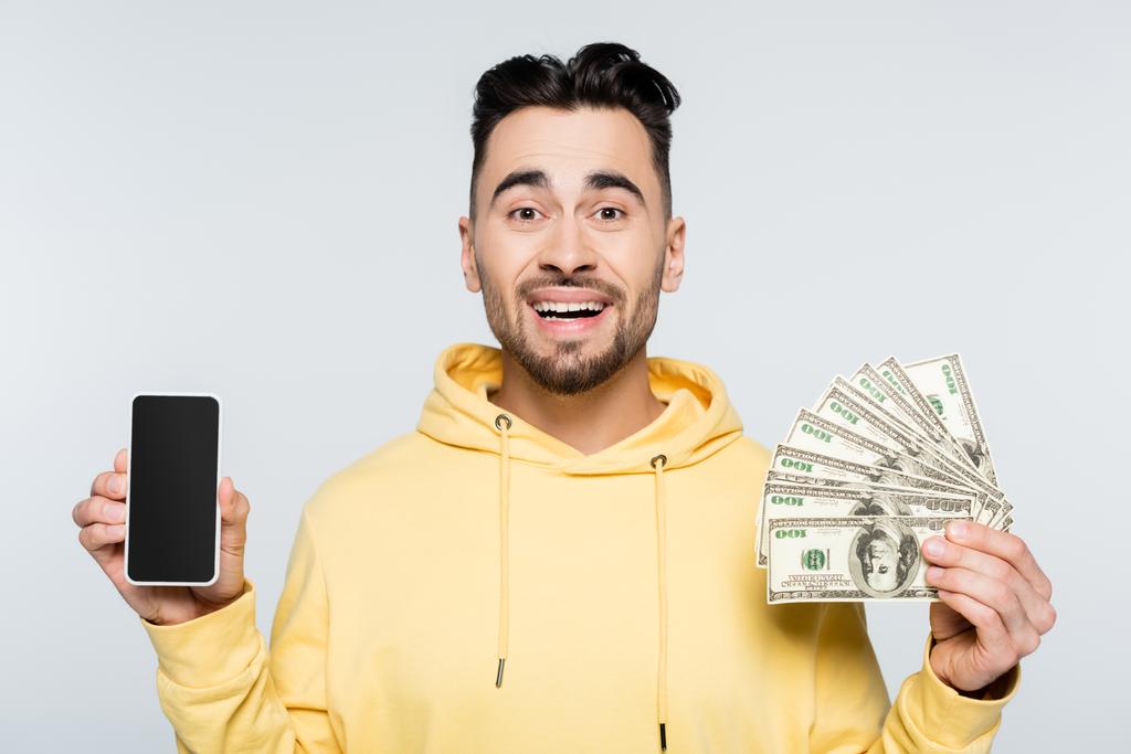cheerful bookmaker holding dollar banknotes and smartphone with blank screen isolated on grey - Photo, image