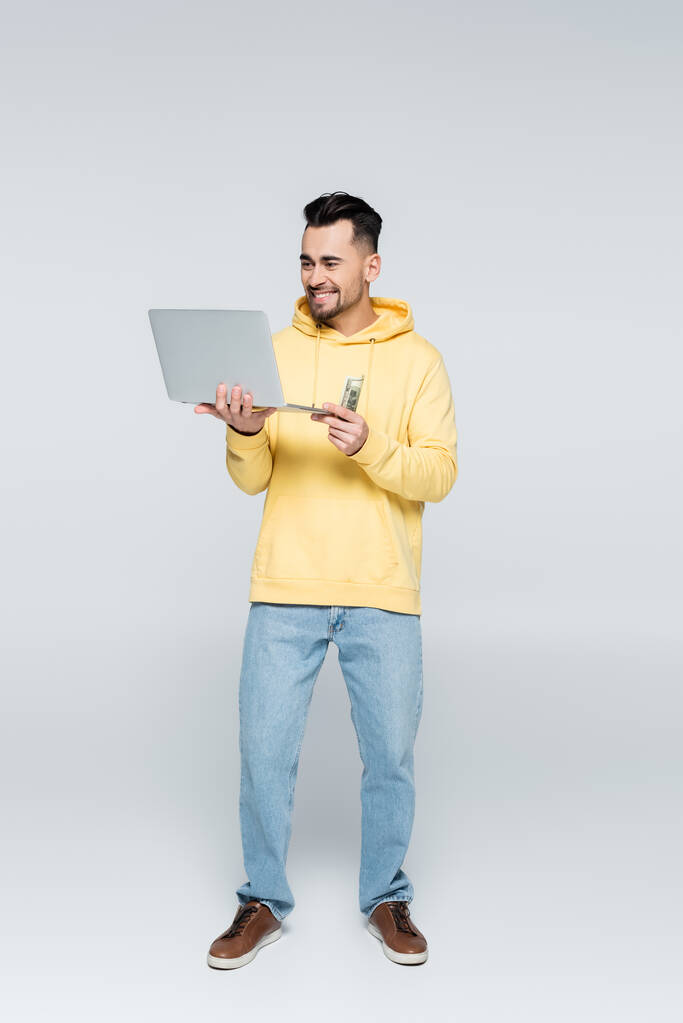 Вид в полный рост счастливого игрока с долларами и ноутбуком на сером - Фото, изображение