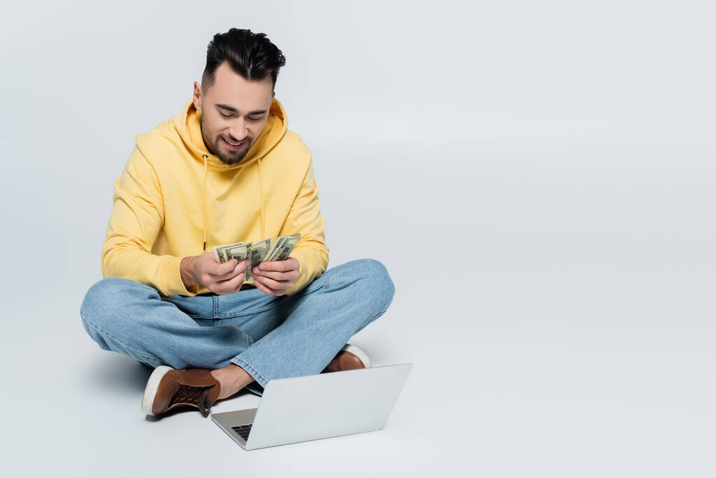pleased man counting dollars while sitting with laptop on grey - 写真・画像