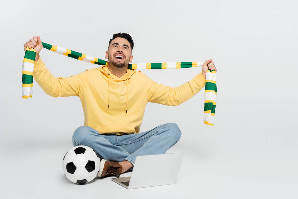 joyful sports fan holding striped scarf while sitting near laptop and soccer ball on grey - 写真・画像