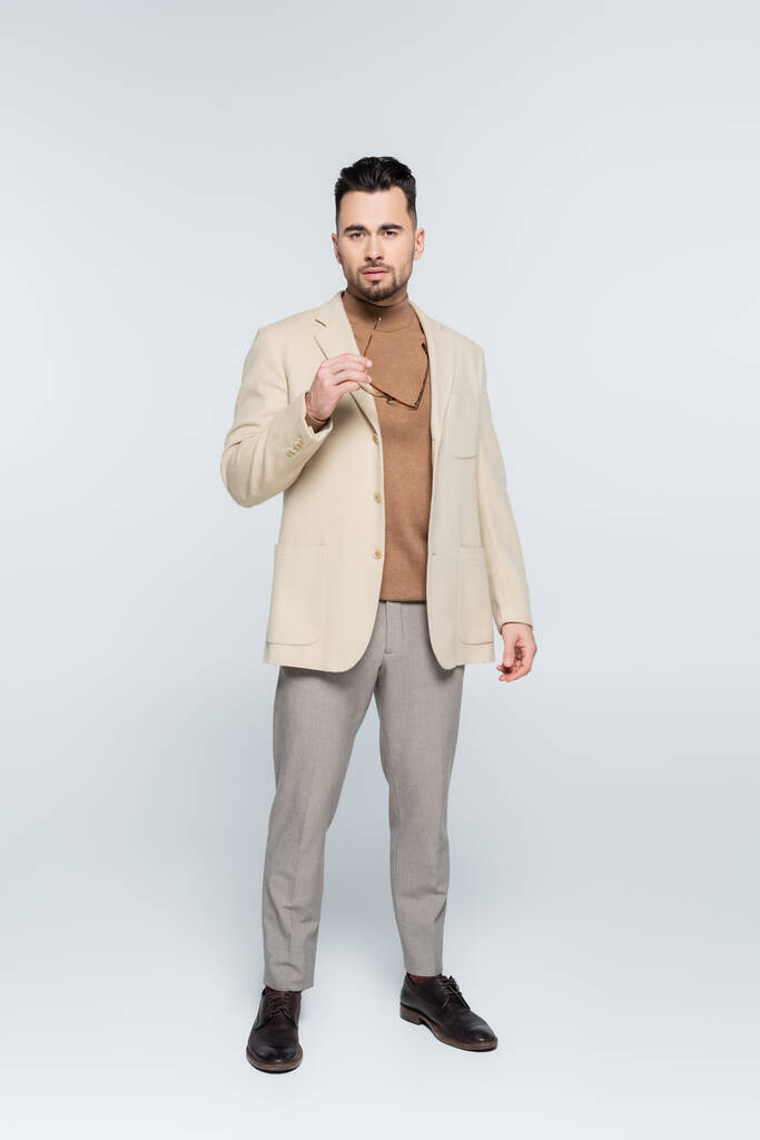 full length of critic in beige blazer and trousers holding eyeglasses on grey - Foto, immagini