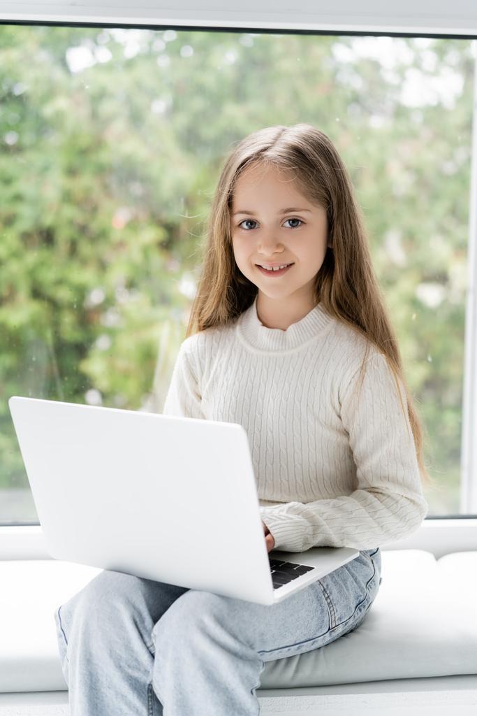 chica feliz mirando a la cámara mientras está sentado cerca de la ventana con el ordenador portátil - Foto, Imagen