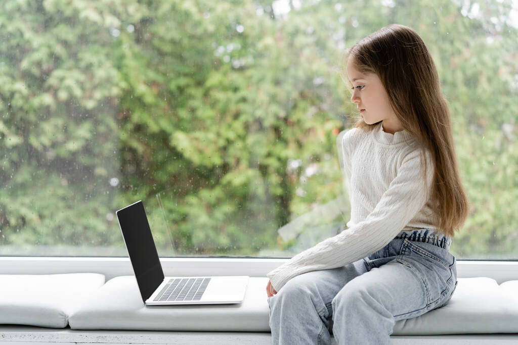 Kız boş ekranlı, dizüstü bilgisayarın yanındaki pencere kenarında oturuyor.  - Fotoğraf, Görsel