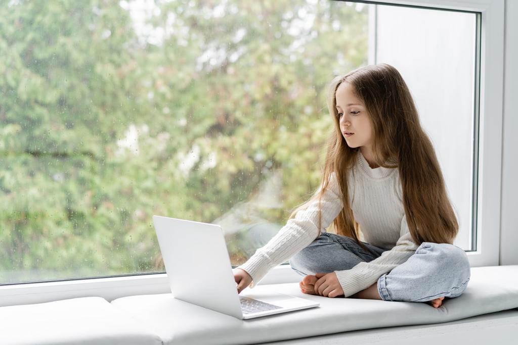 girl sitting on windowsill with crossed legs and using laptop during online lesson - 写真・画像