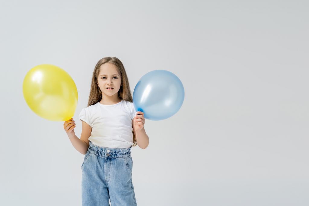 fille heureuse avec des ballons bleus et jaunes regardant la caméra isolée sur gris - Photo, image