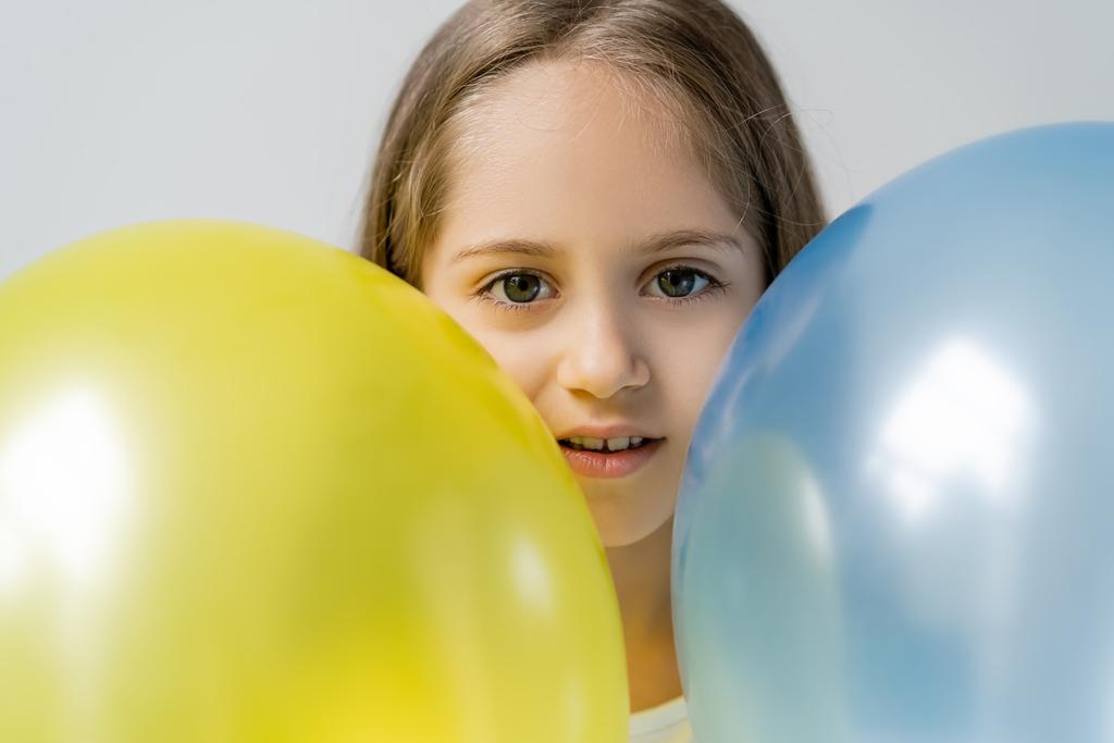 menina patriótica olhando para a câmera perto de balões azuis e amarelos isolados em cinza - Foto, Imagem