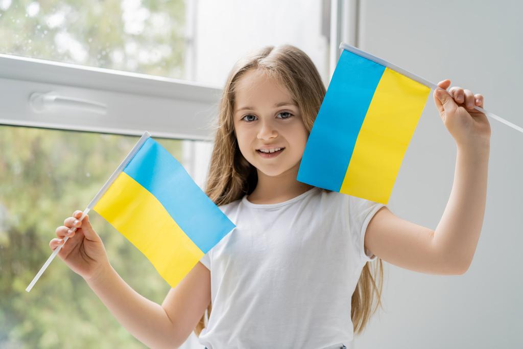 Девушка с маленькими украинскими флагами улыбается у окна дома - Фото, изображение