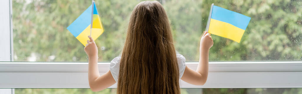 visão traseira da menina com cabelos longos segurando pequenas bandeiras ucranianas perto da janela, bandeira - Foto, Imagem