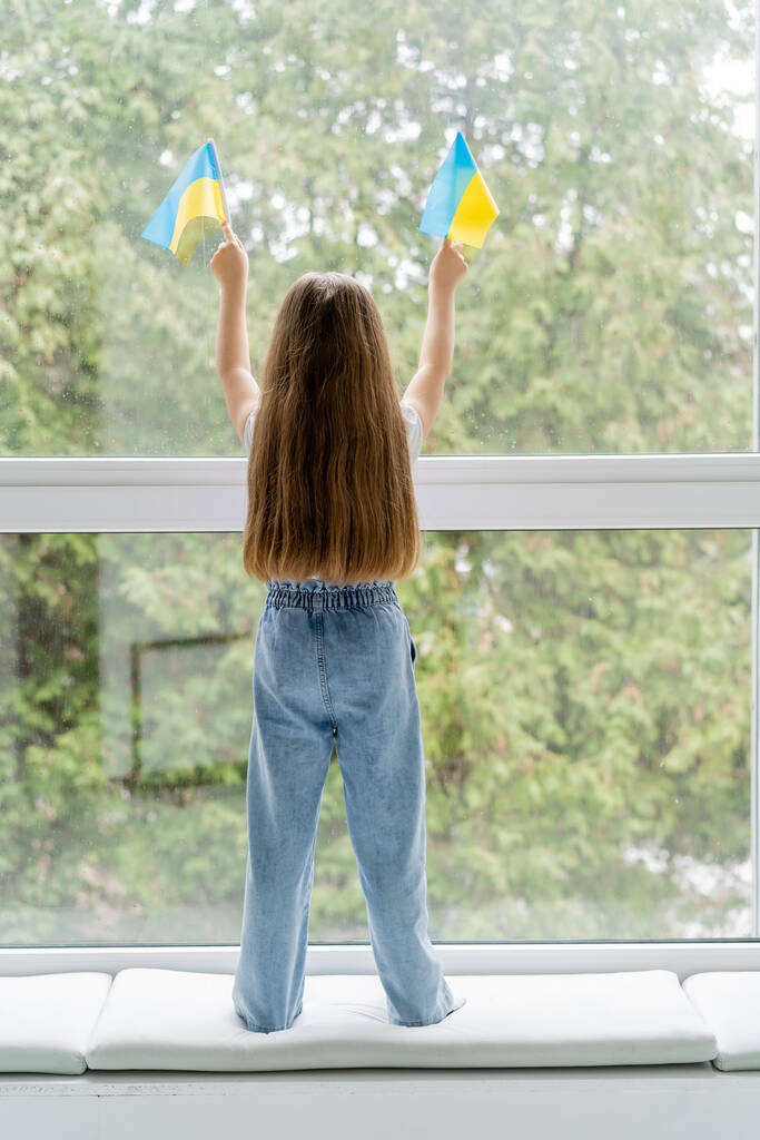 πλήρες μήκος του κοριτσιού με μακριά μαλλιά στέκεται με μικρές ουκρανικές σημαίες κοντά στο παράθυρο, πίσω όψη - Φωτογραφία, εικόνα