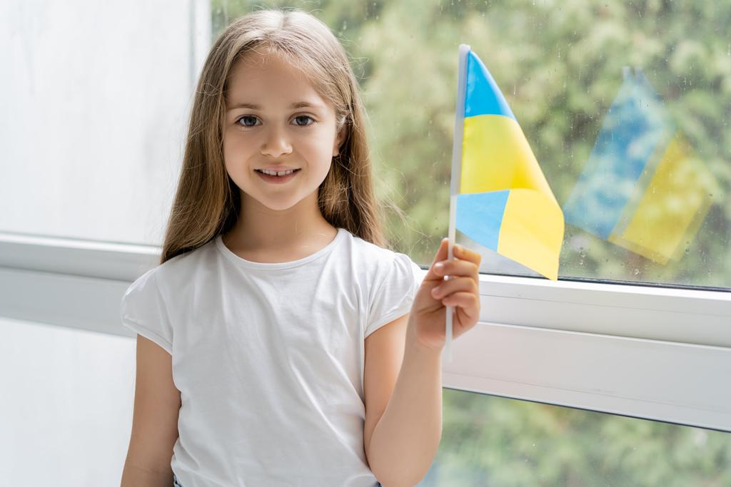 χαρούμενο κορίτσι με μικρή ουκρανική σημαία κοιτάζοντας κάμερα κοντά στο παράθυρο στο σπίτι - Φωτογραφία, εικόνα