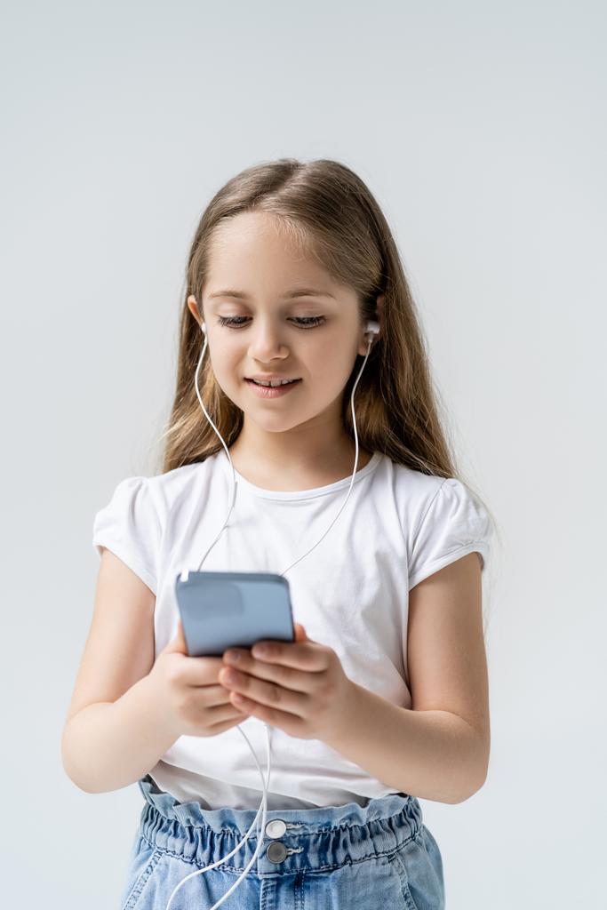 glimlachend meisje in oortelefoons met behulp van mobiele telefoon geïsoleerd op grijs - Foto, afbeelding