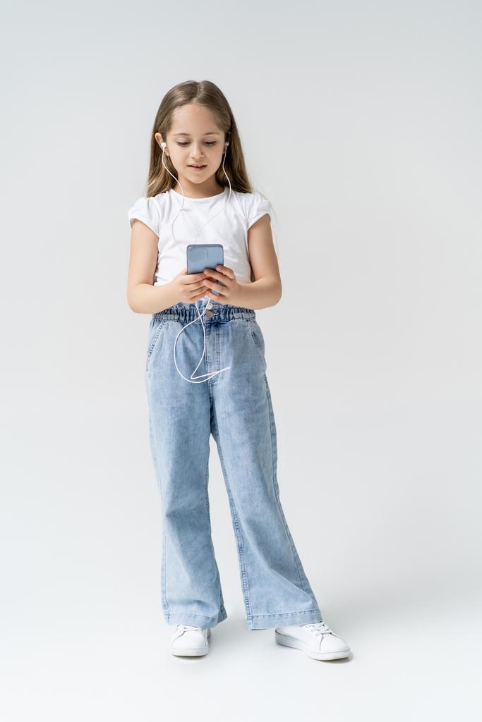 vista a figura intera di ragazza in jeans e auricolari utilizzando smartphone su grigio - Foto, immagini
