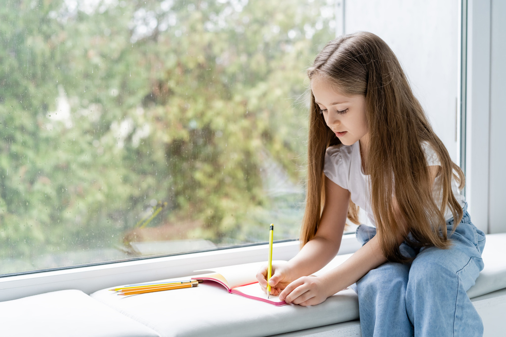 menina com cabelos longos fazendo lição de casa no peitoril da janela perto de vidro com gotas de chuva - Foto, Imagem