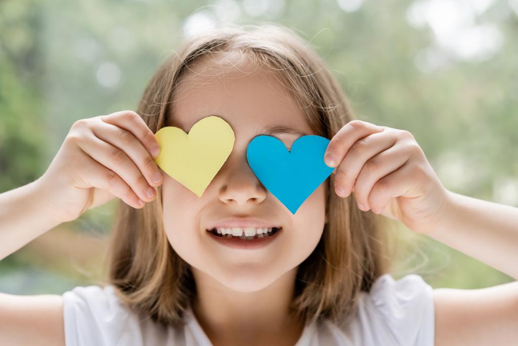 cheerful ukrainian girl obscuring face with blue and yellow paper hearts - 写真・画像
