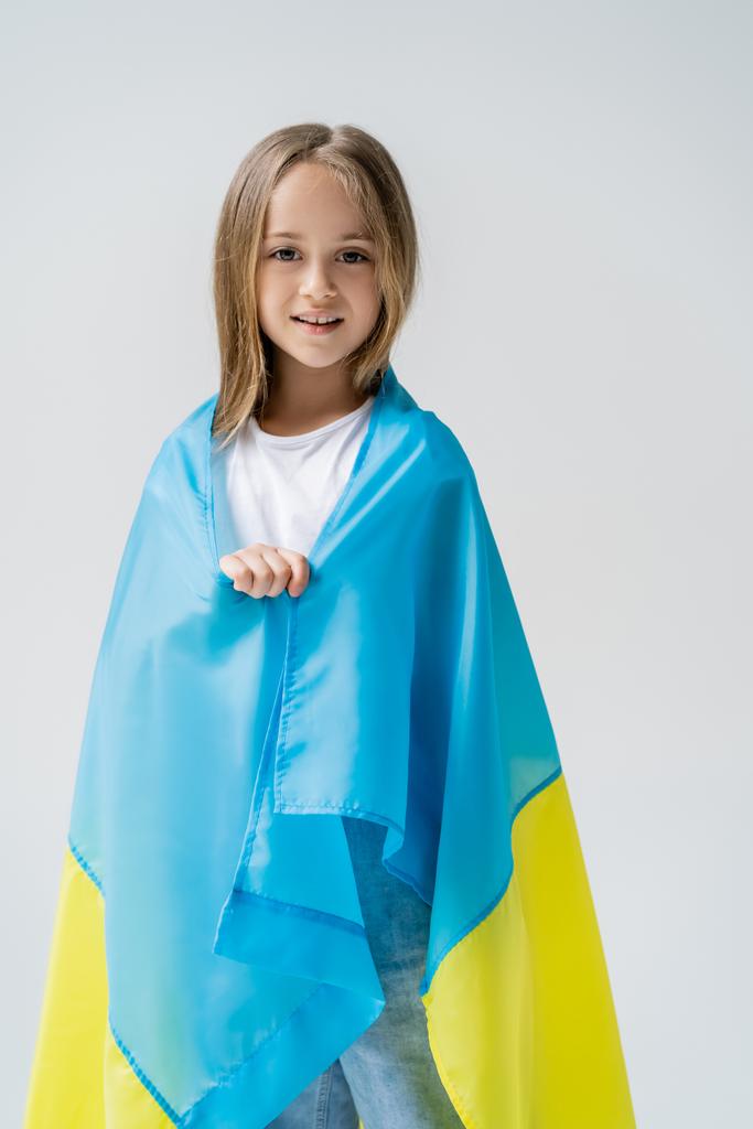 chica patriótica cubierta con bandera ucraniana sonriendo a la cámara aislada en gris - Foto, Imagen