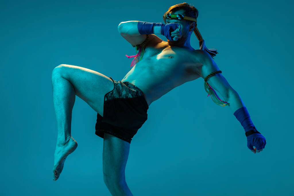 Kolenní kop. Dynamický portrét profesionálního thajského boxera cvičícího izolovaně na modrém pozadí studia. Sport, muay thai, soutěž, koncept klubu rváčů - Fotografie, Obrázek