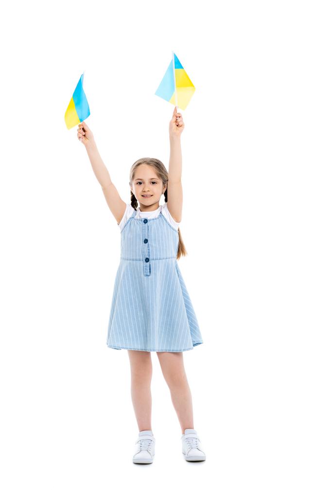 Ganzkörperansicht des Mädchens, das mit ukrainischen Flaggen in erhobenen Händen auf weiß steht - Foto, Bild
