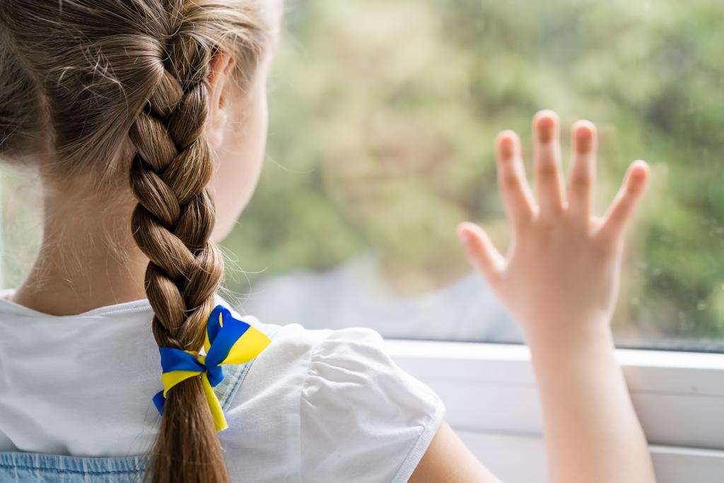 vista posteriore della ragazza con nastro blu e giallo sulla treccia in piedi vicino alla finestra sfocata - Foto, immagini