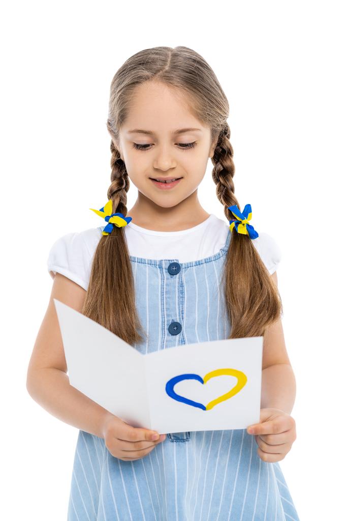 girl with blue and yellow ribbons on braids reading card with heart sign isolated on white - Photo, Image