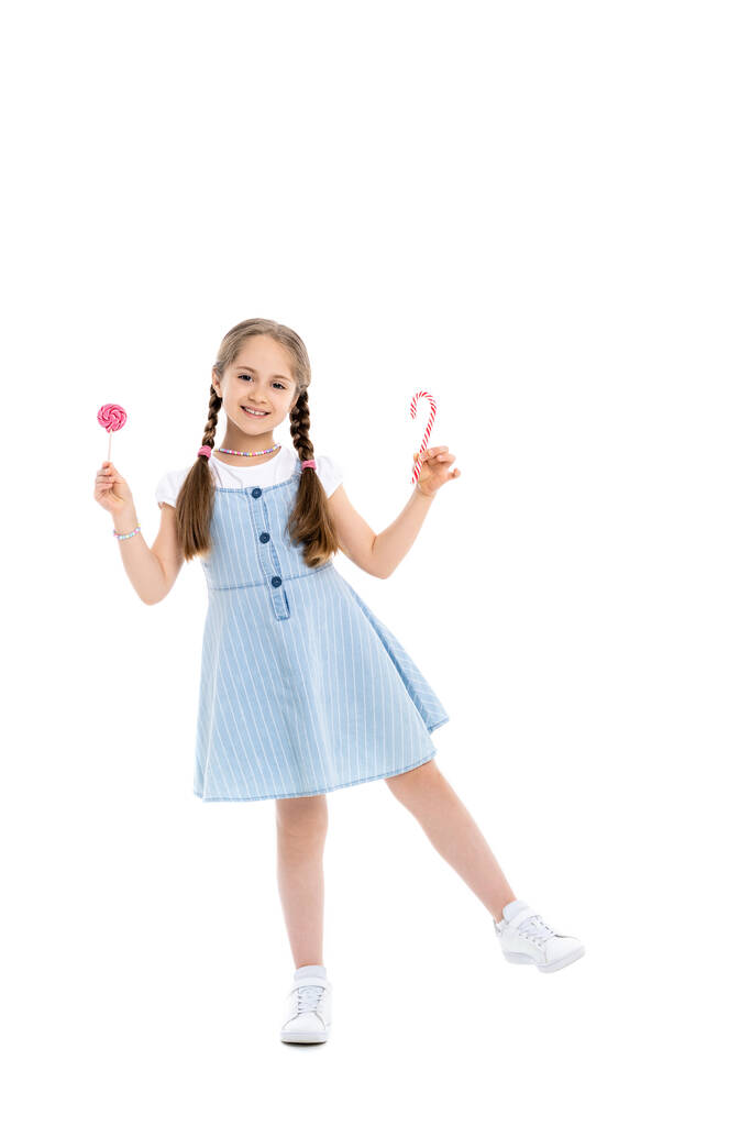 full length view of cheerful girl with candy cane and lollipop on white - Photo, Image