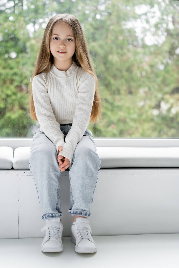 visão de comprimento total da menina em jeans sentado no peitoril da janela e olhando para a câmera - Foto, Imagem