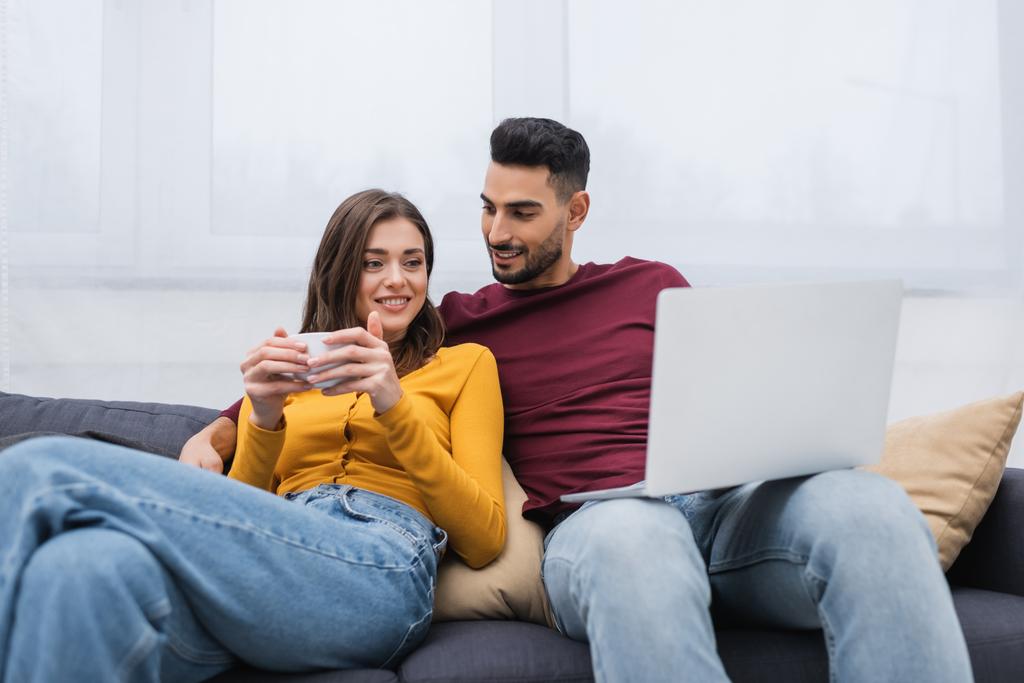 Cheerful woman holding cup of coffee near arabian boyfriend with laptop on couch  - Foto, Imagem