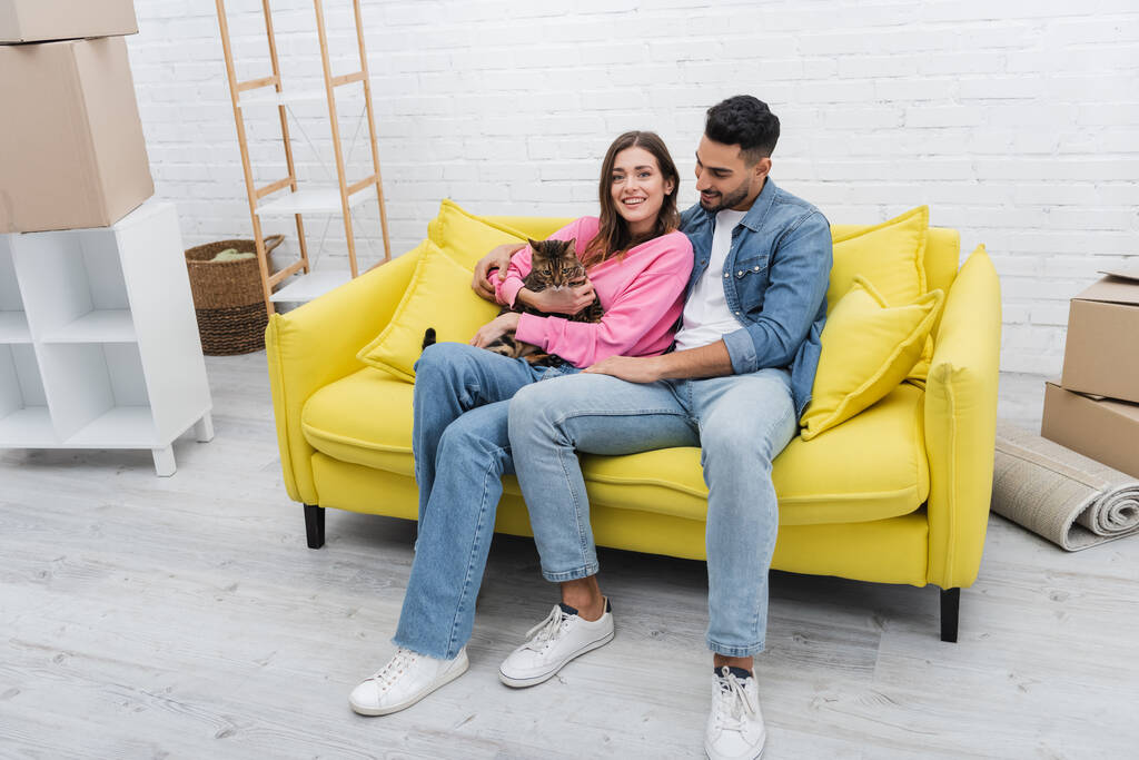Happy woman smiling at camera while holding bengal cat near muslim boyfriend on couch  - Photo, Image