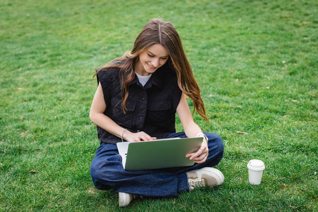 χαρούμενη νεαρή γυναίκα που χρησιμοποιεί φορητό υπολογιστή ενώ κάθεται στο γκαζόν - Φωτογραφία, εικόνα