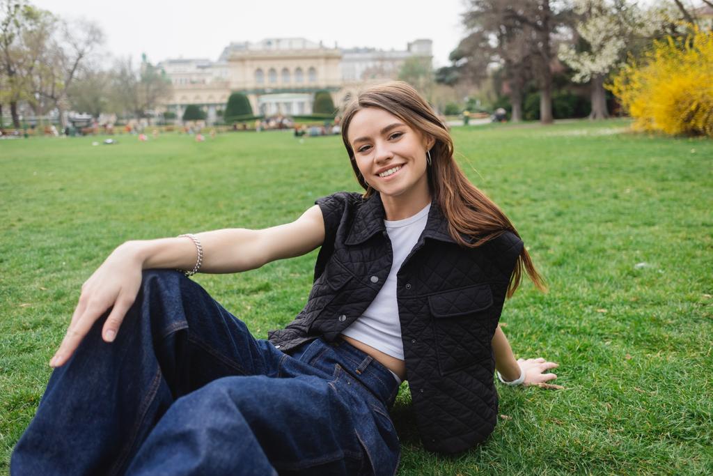 joyful young woman in sleeveless jacket sitting on lawn  - 写真・画像