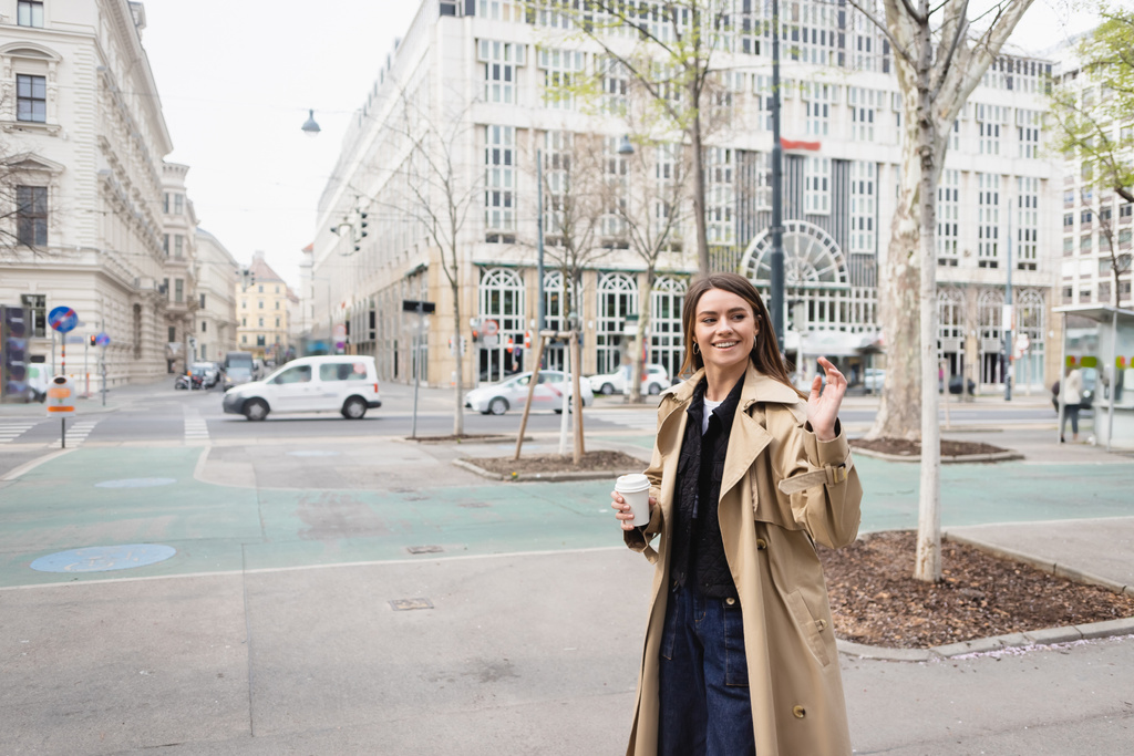 glückliche junge Frau in stylischem Trenchcoat, die mit Coffee to go durch die europäische Stadt läuft und winkt - Foto, Bild