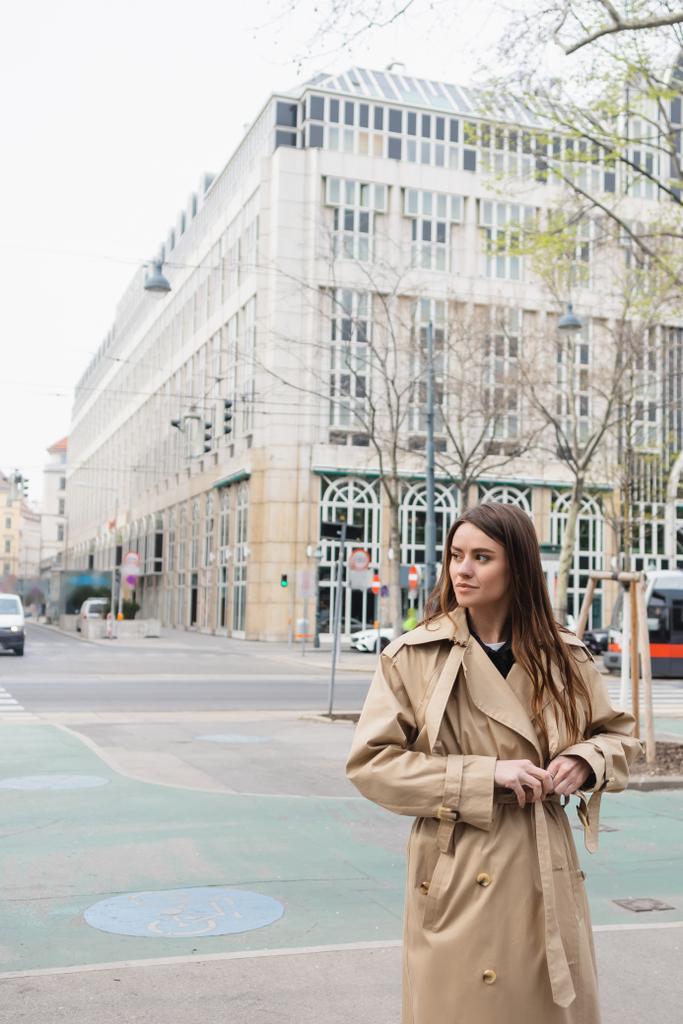 молодая женщина натягивает пояс на стильное пальто на улице европейского города - Фото, изображение