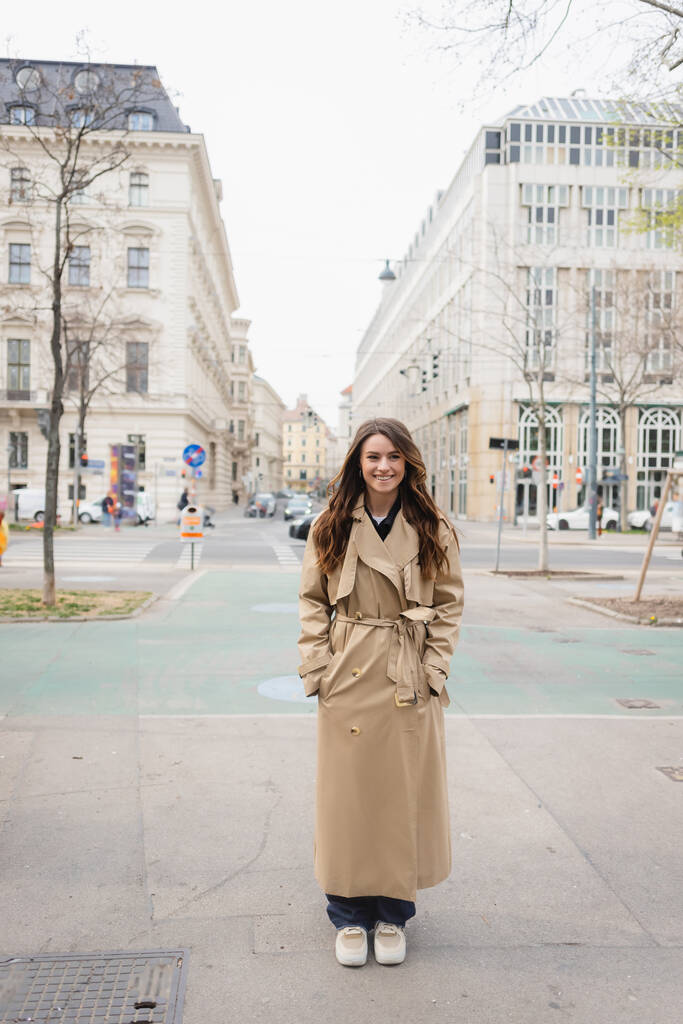 comprimento total da jovem mulher feliz em casaco de trincheira de pé com as mãos em bolsos na rua da cidade europeia - Foto, Imagem